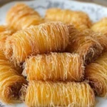 A plate of golden, crispy kataifi pastries, tightly wrapped in fine strands of shredded phyllo dough, arranged in a pile on an ornate white and gray plate.