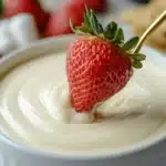 A bowl of creamy cheesecake fondue with a strawberry dipped in it, dripping with rich, velvety sauce.